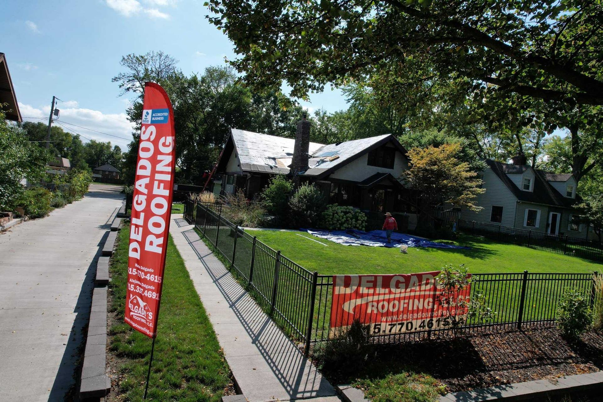 Delgra roofing signs on a sidewalk beside a house and lawn in a sunny neighborhood