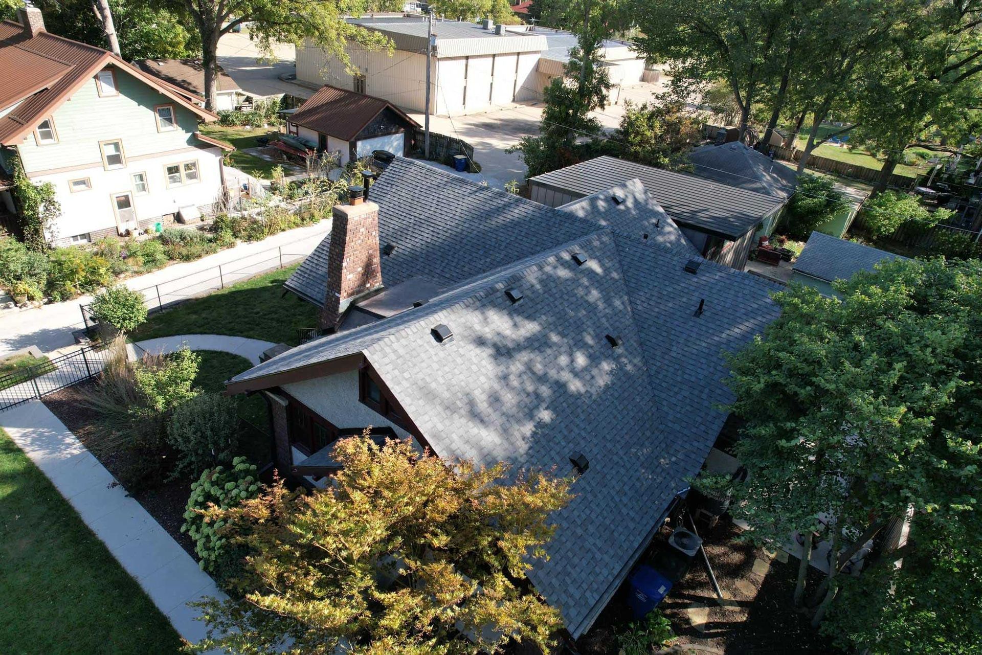 Aerial view of a house with a dark roof surrounded by trees and neighboring homes.