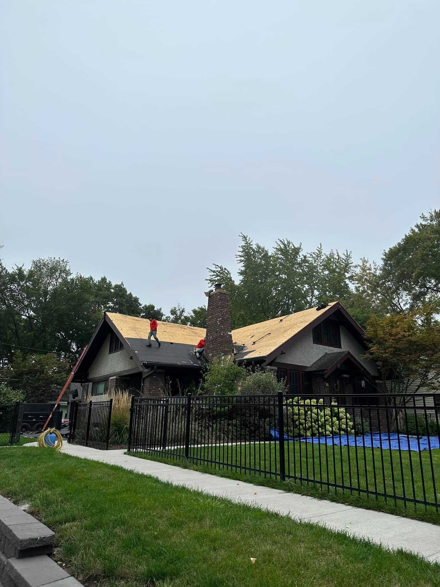 House with two workers repairing the roof, behind a black fence and sidewalk under a cloudy sky