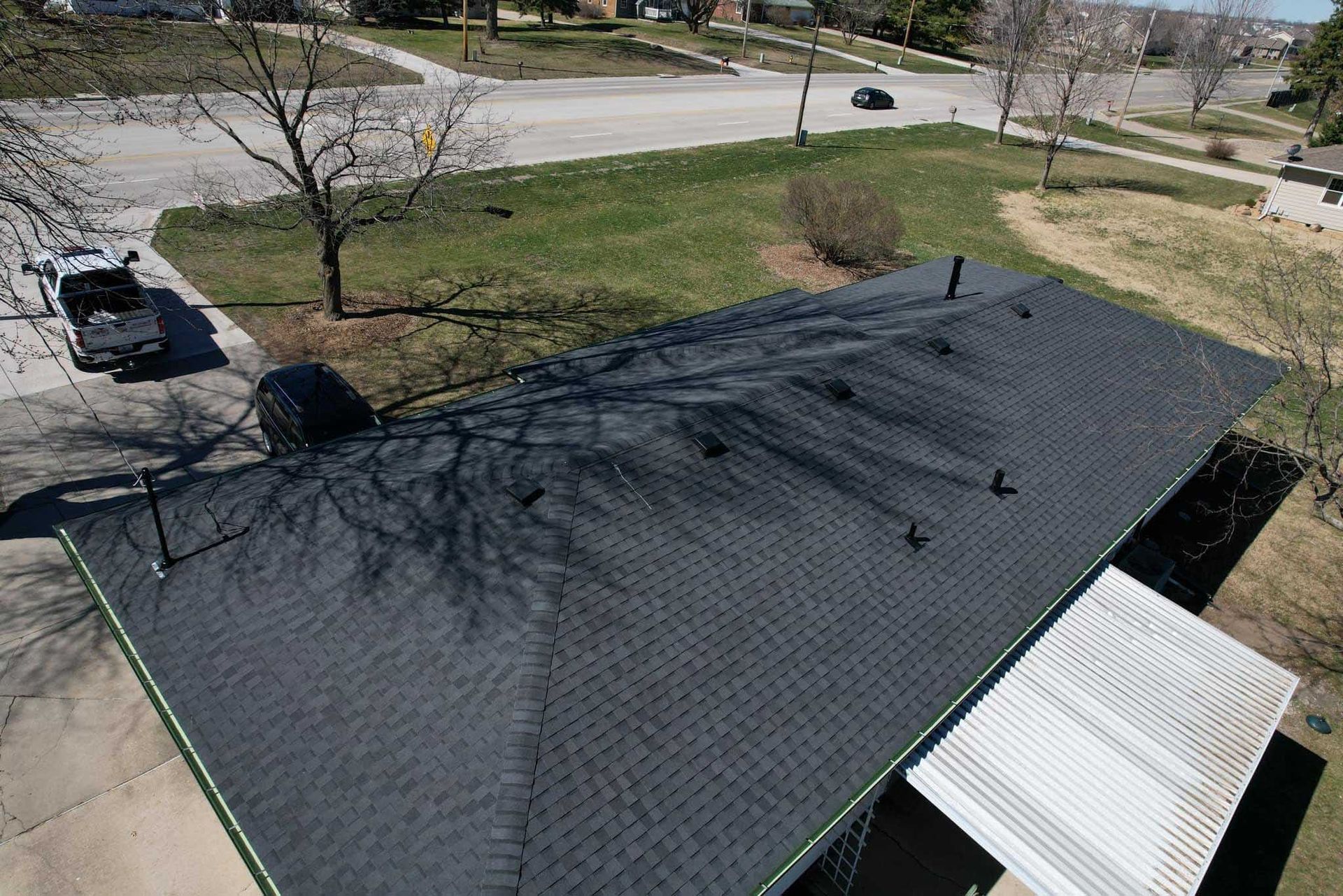 Aerial view of a dark shingled roof with trees and a street in the background.