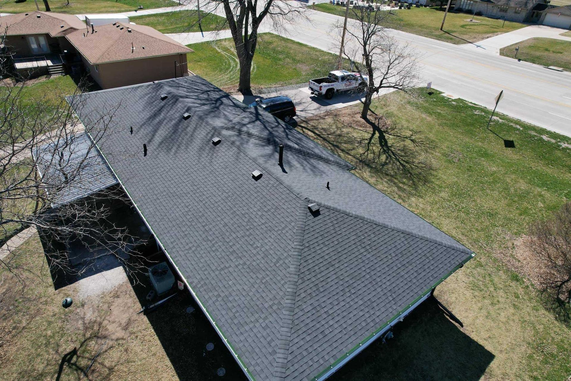 Aerial view of a gray roof with a tree casting shadows beside a grassy yard and street.