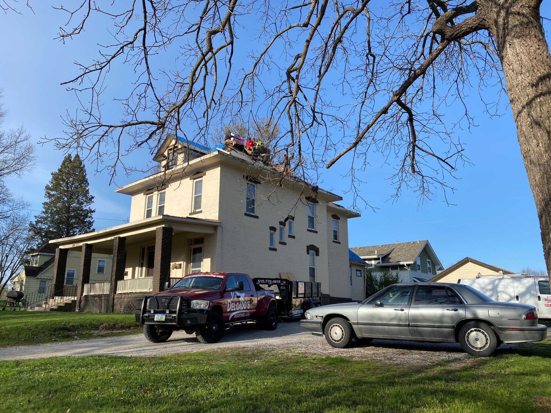 Two-story beige house with parked cars and workers on the roof under a blue sky