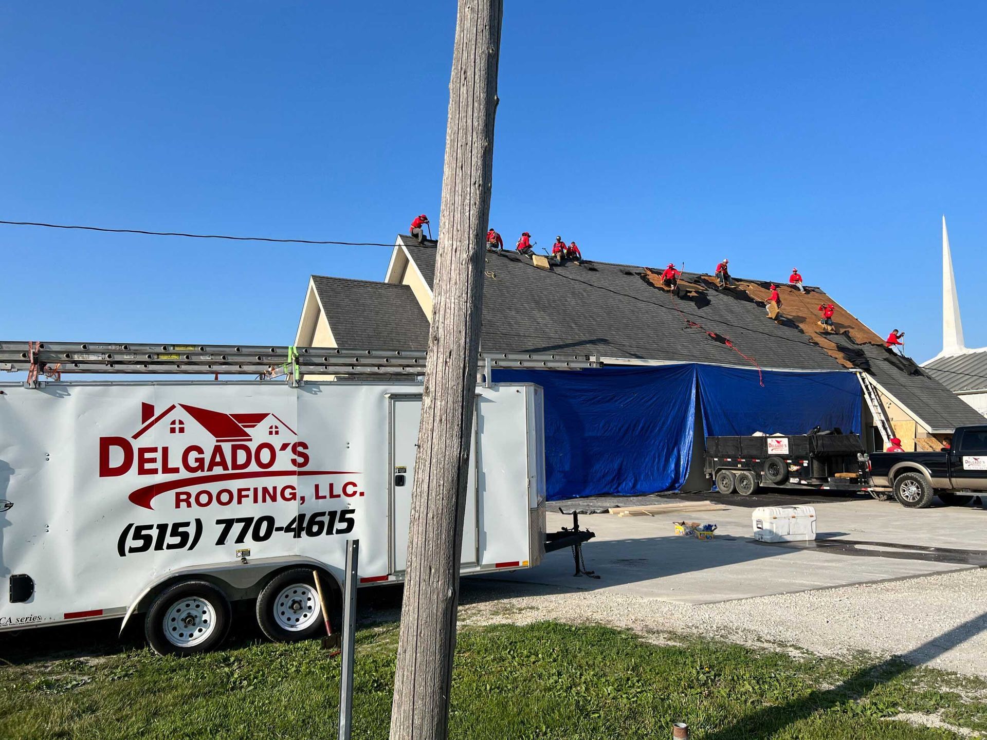 Roofers working on a house with a white Delgado's Roofing trailer in front under a blue sky