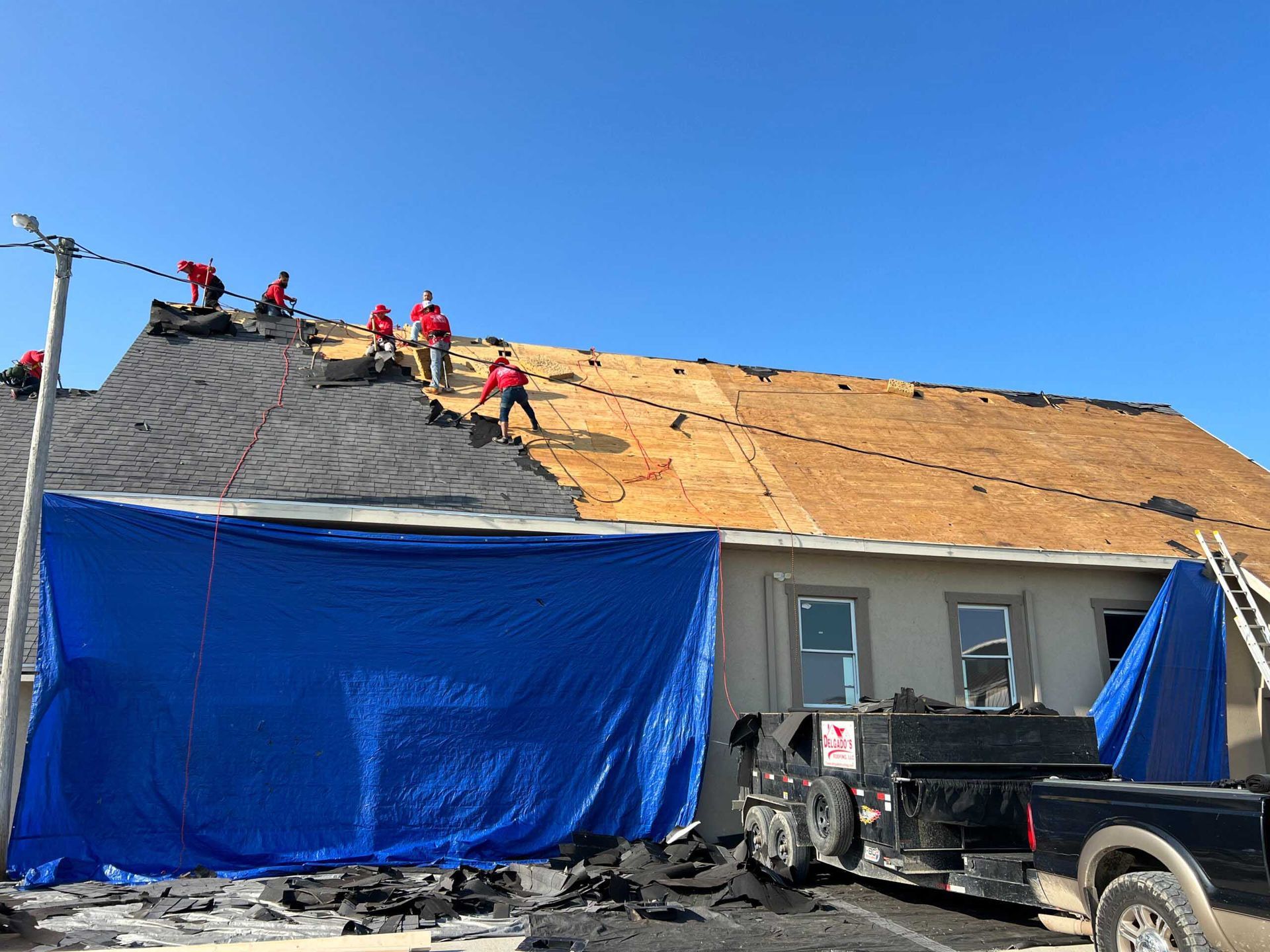Workers reroofing a house, with blue tarp coverings and a black pickup truck in front.