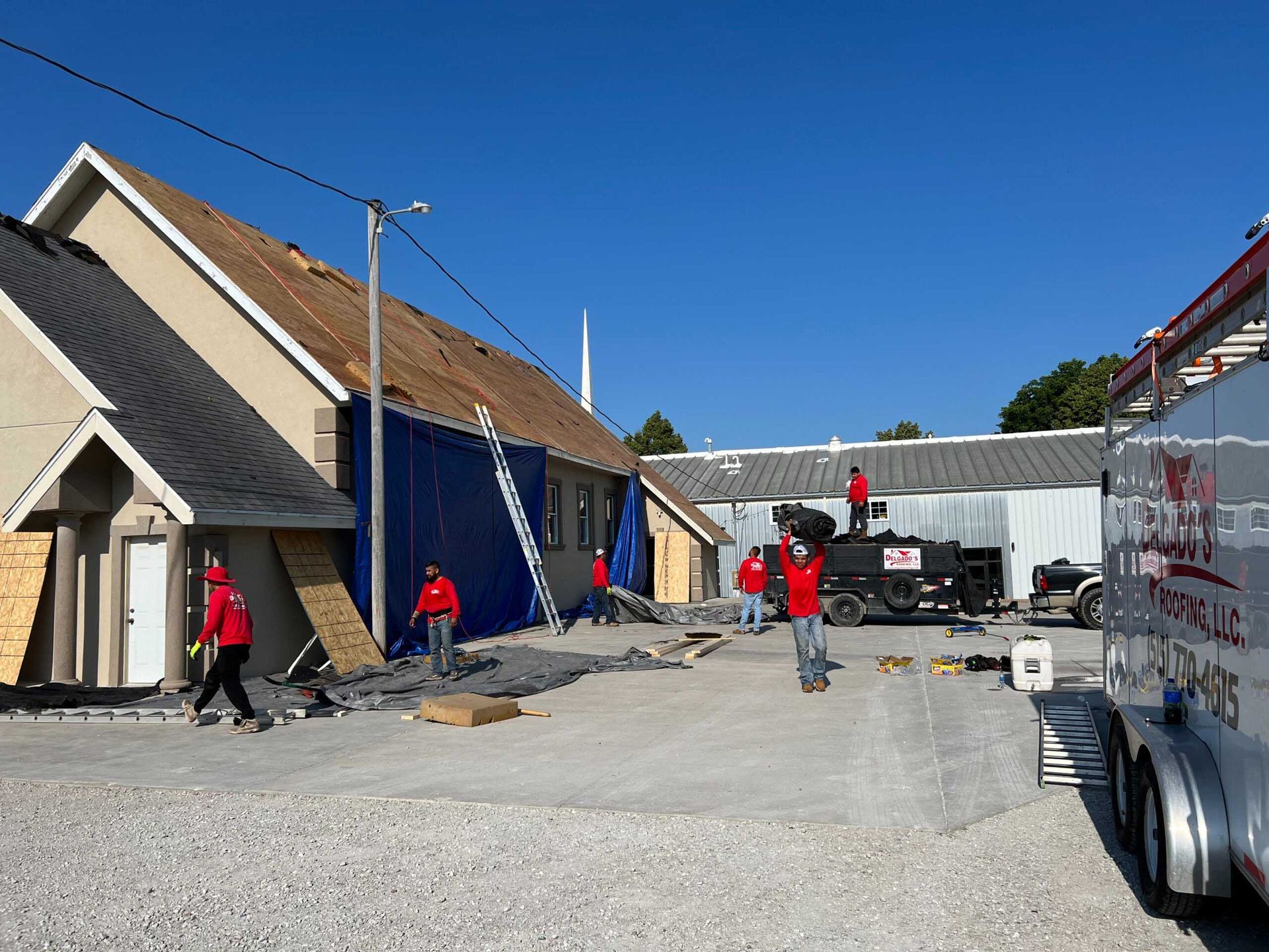 Workers on a construction site beside blue buildings and a parked trailer under a clear sky