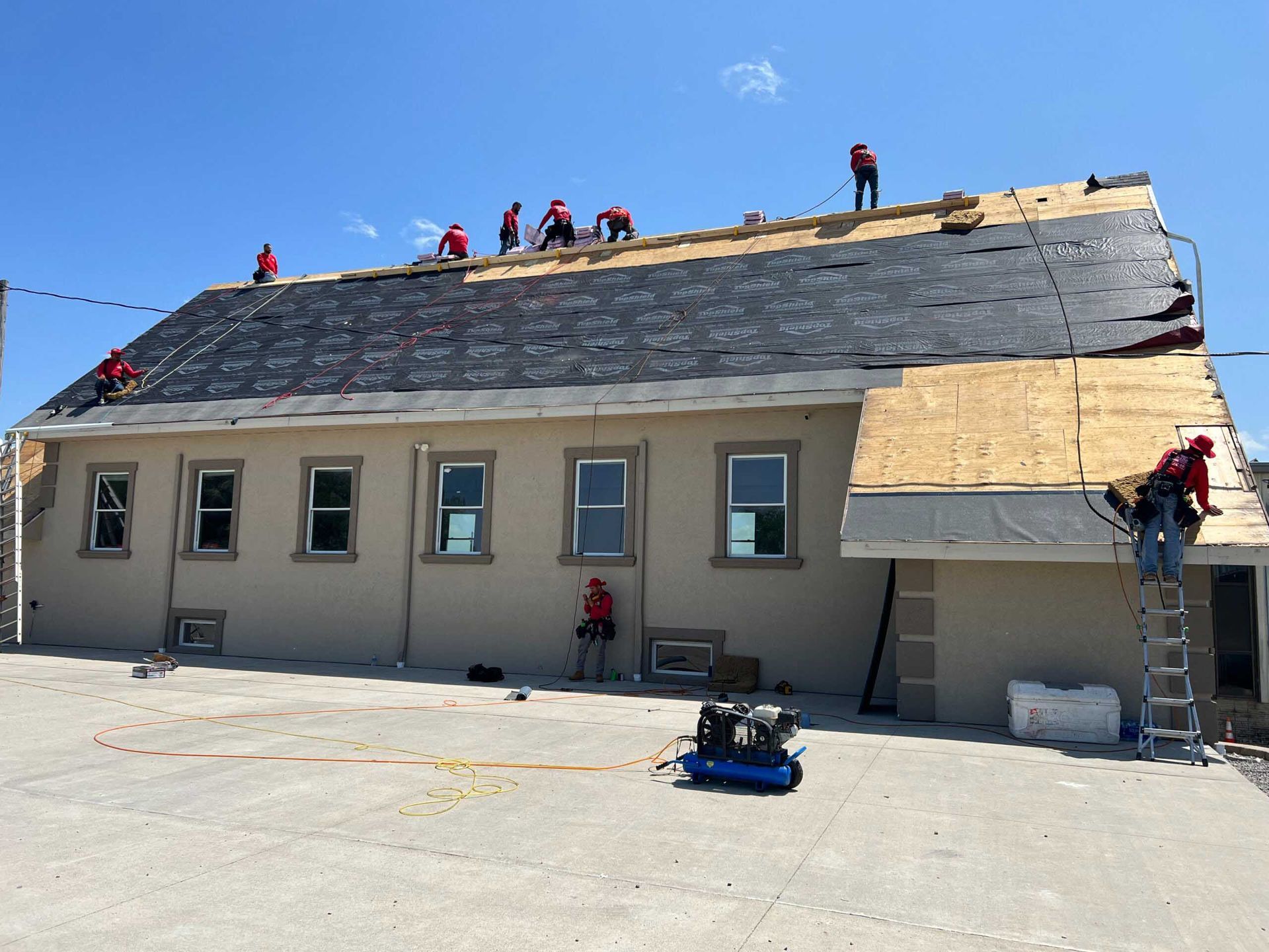 Workers reroofing a beige building with gray shingles and a ladder under a clear blue sky