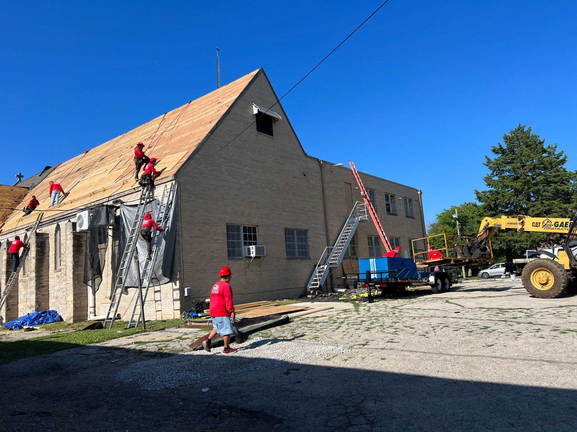 Workers on ladders repairing a brown building roof beside a blue lift and yellow loader.