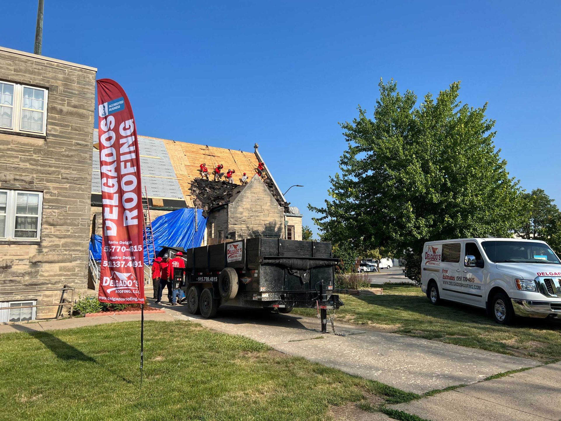 Workers beside a black machine and white van at a stone building under a bright blue sky