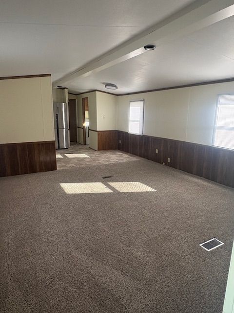 Empty living room with brown carpet, wood paneling, and a glimpse of the kitchen.