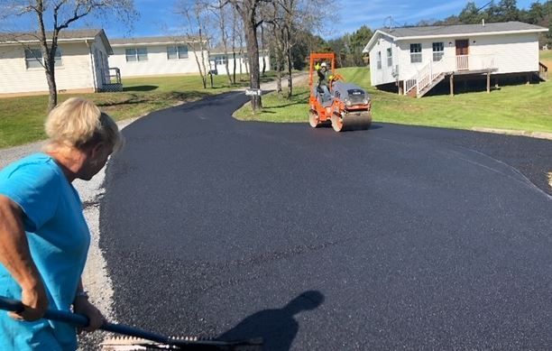 A woman in a blue shirt is standing on a road while a roller is rolling asphalt.