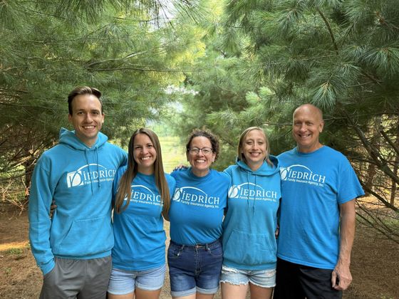 Five people in blue shirts standing outdoors among trees, smiling at the camera.