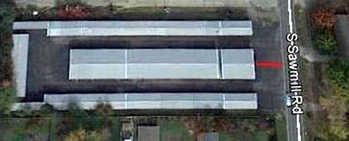 Aerial view of a long gray-roofed building beside a road and trees