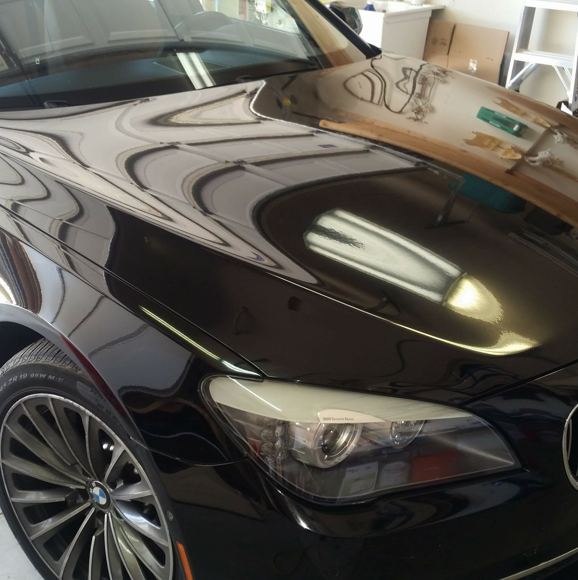 Shiny black sports car hood and headlight in a garage, reflecting overhead lights