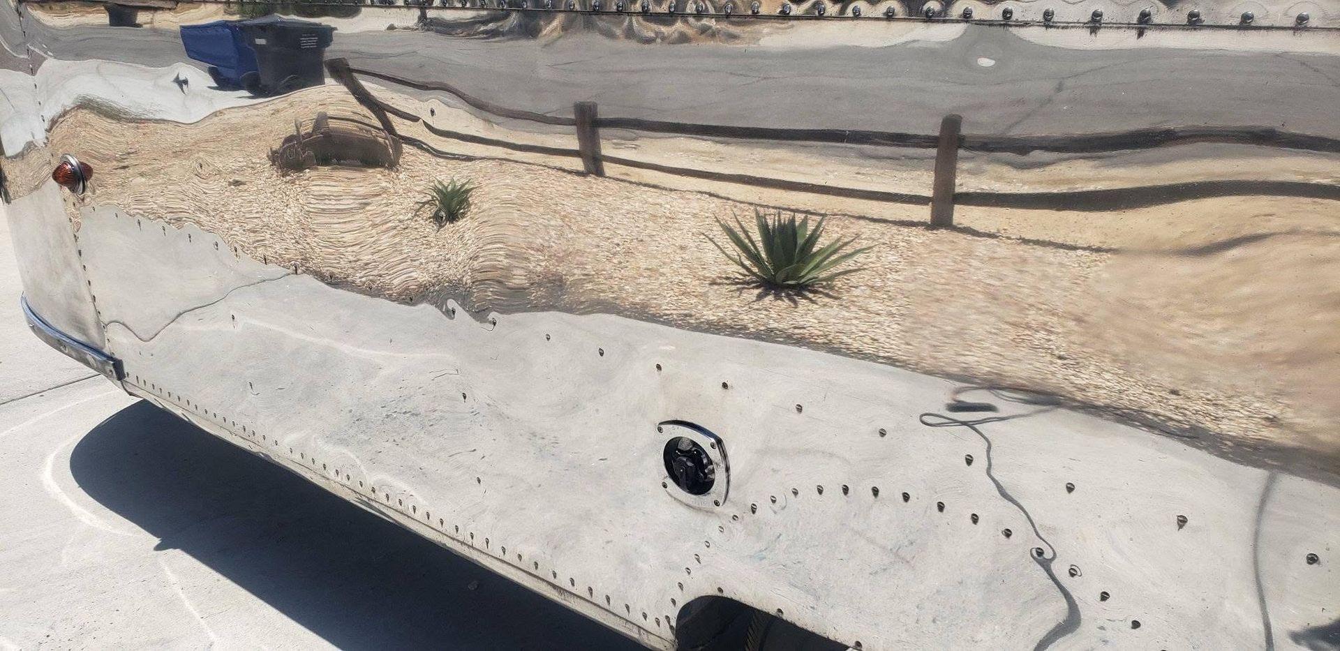 Concrete roadside curb beside a dry landscaped area with gravel, small plants, and a dark drain opening