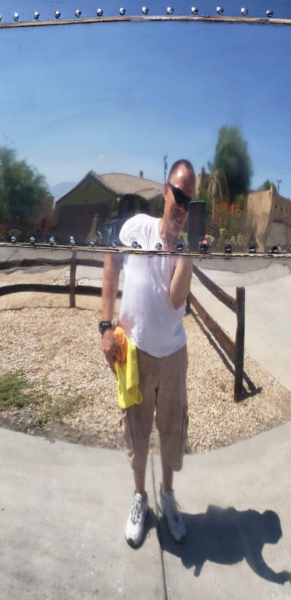 Person standing outdoors on a sunny dirt path, holding a yellow bag, with a fence and hills in the background.