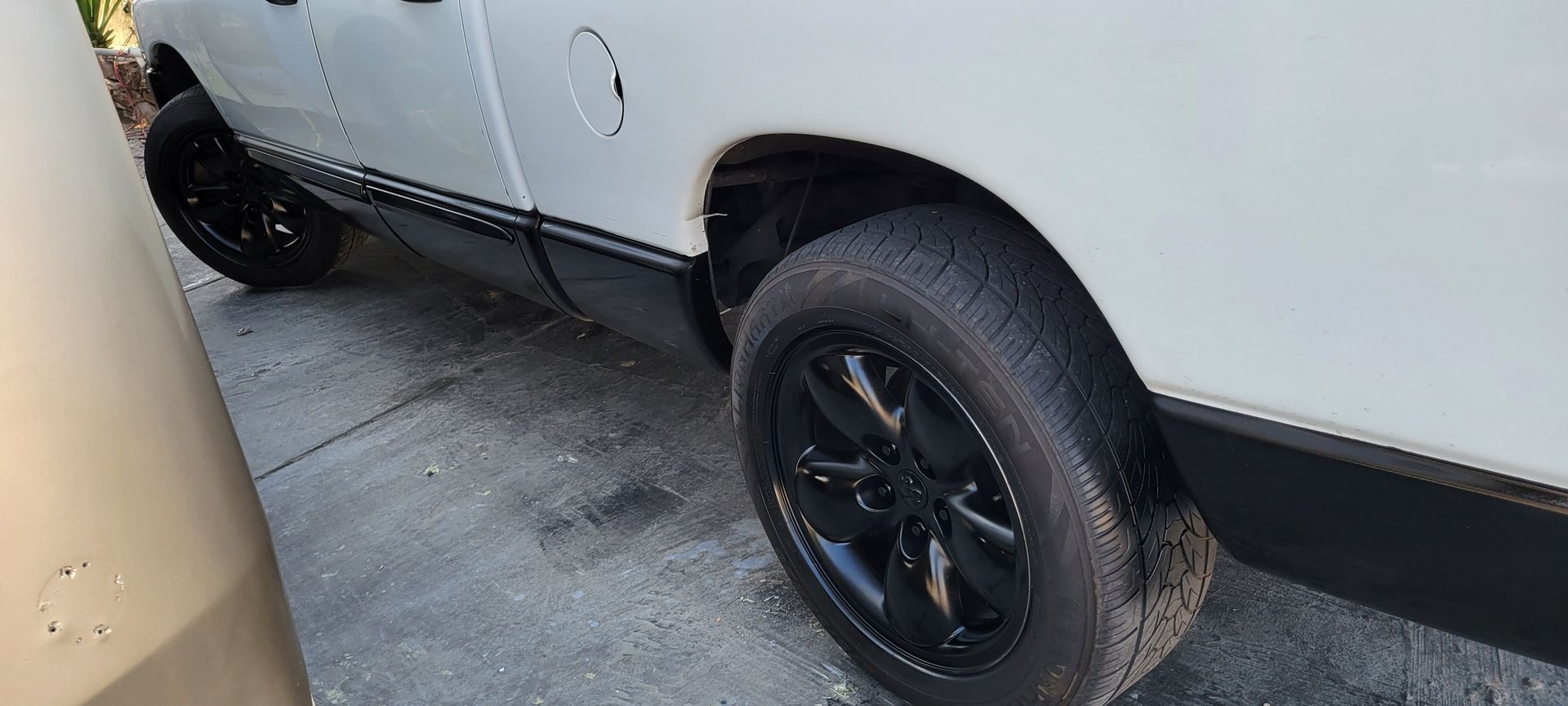 White SUV with black wheels parked in a narrow driveway, close to a beige vehicle.