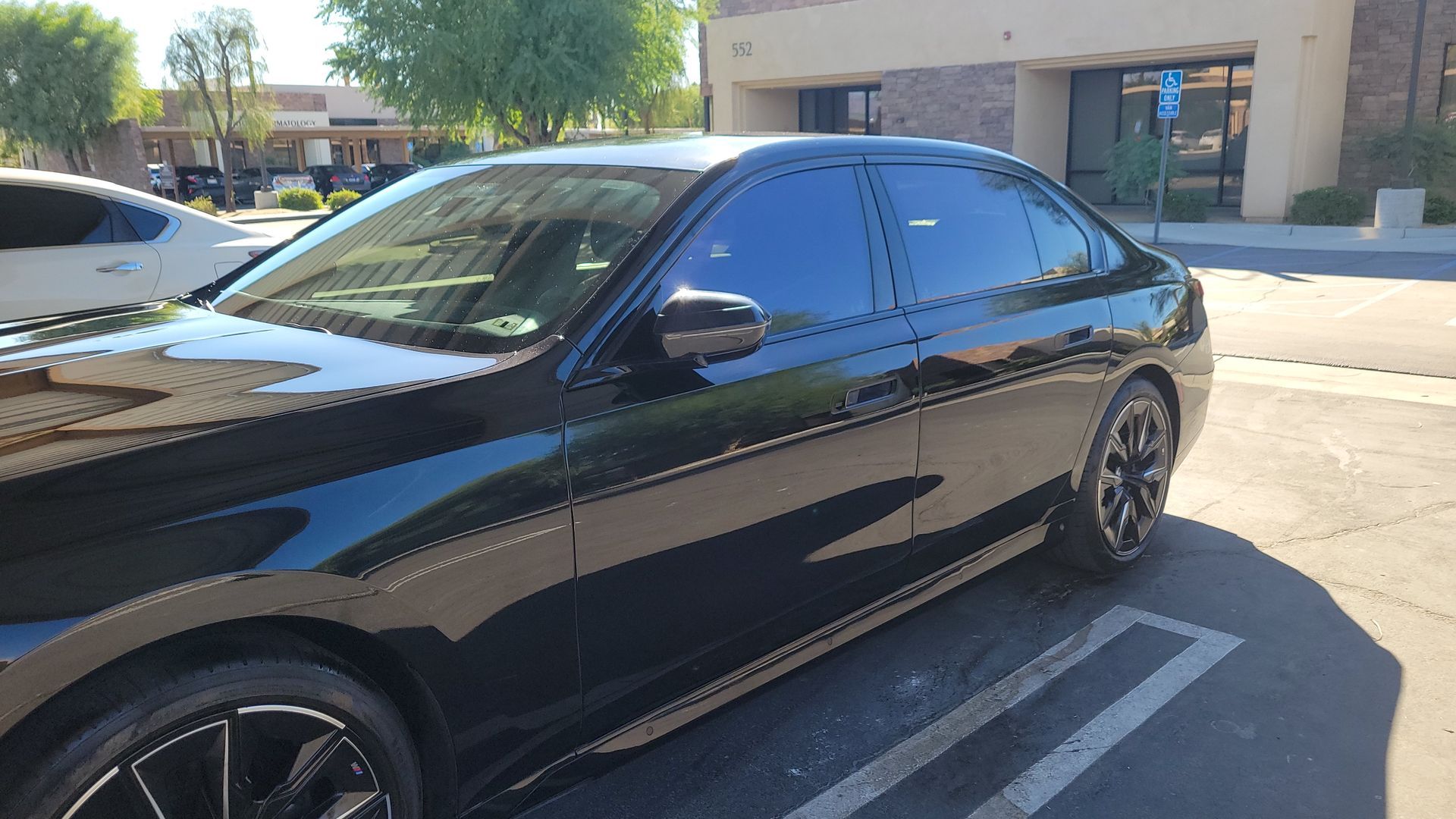 Black sedan parked in a sunlit lot beside a building, viewed from the side.