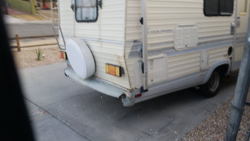 White camper trailer parked on a concrete driveway, rear corner visible with spare tire cover and small window.