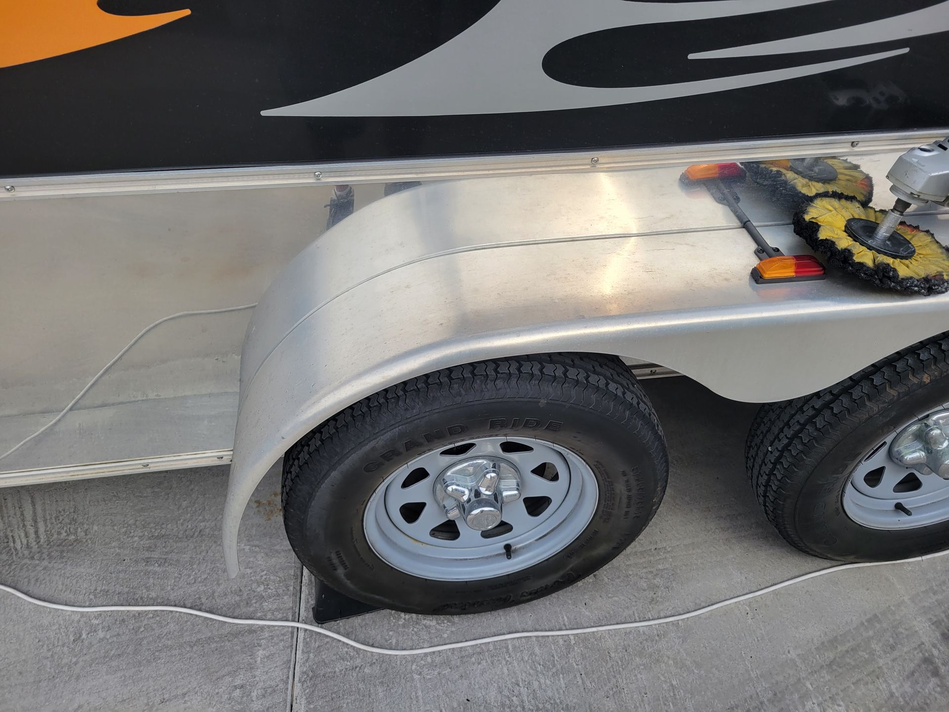Close-up of a silver trailer’s wheel and fender on pavement, with a black and orange logo above.