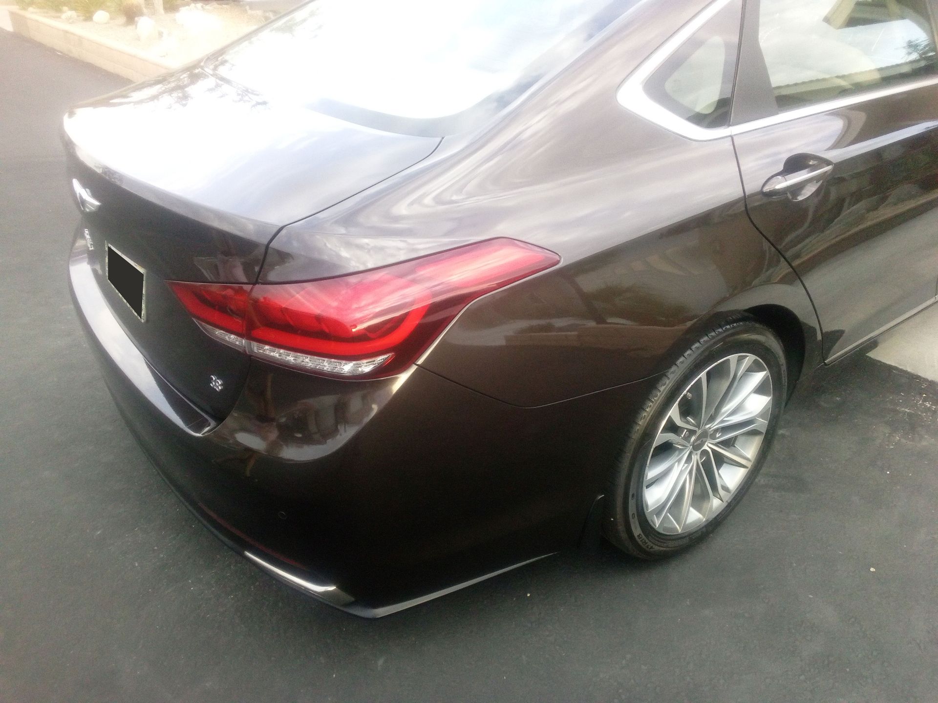 Rear view of a dark brown sedan parked on pavement, showing the taillight and rear wheel.