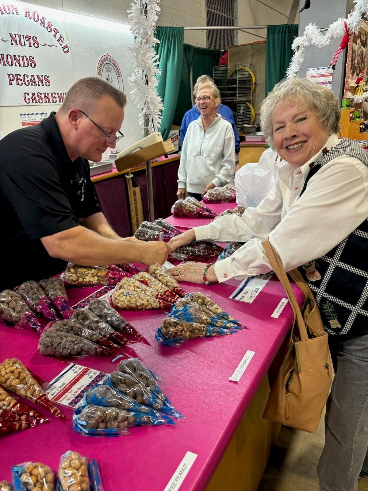 Vendor smiling behind a pink table of packaged goods at a market stall while a man handles items nearby