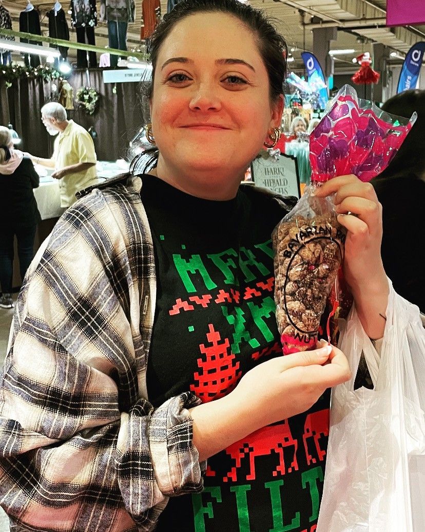 Person smiling in a market, holding a wrapped bouquet and a decorated item, wearing a plaid shawl and festive shirt
