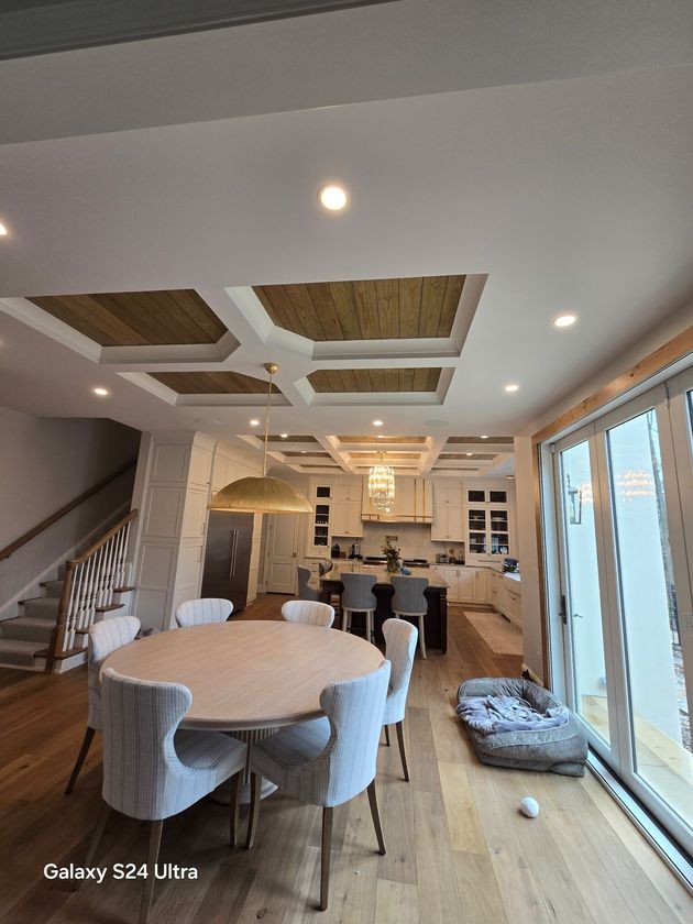 Open-concept living room and kitchen with round dining table, white chairs, wood floors, and large windows.