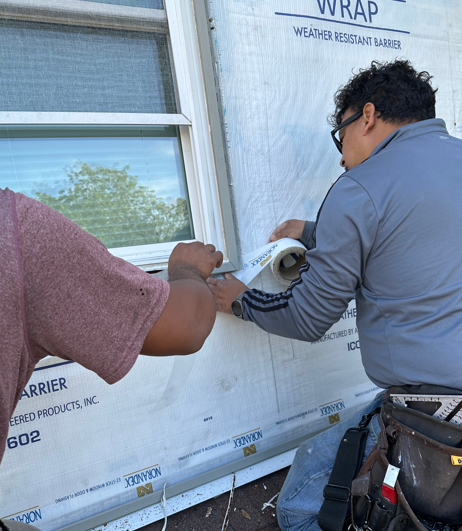 Two workers install window wrap on a house exterior, using a roller and smoothing the material.