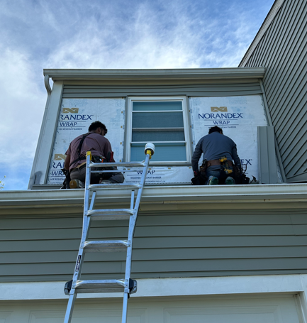 Workers on a ladder installing siding on an upper-story house window under a blue sky