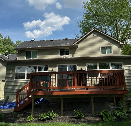 Two-story beige house with a wooden deck and stairs in a green backyard under a blue sky