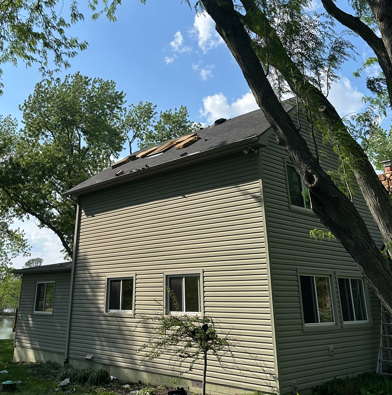 Two-story beige house with damaged roof and trees overhead on a sunny day