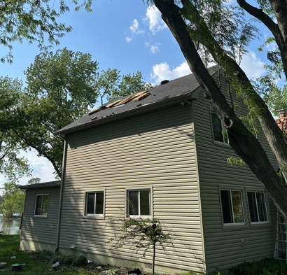 Two-story beige house with damaged roof under tall trees on a sunny day