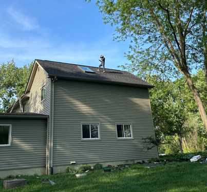 Gray two-story house with a sloped roof, skylight, and trees in a grassy yard under a blue sky
