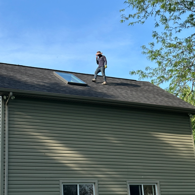 Worker on a rooftop beside a skylight, standing on a gray shingled house under a clear blue sky