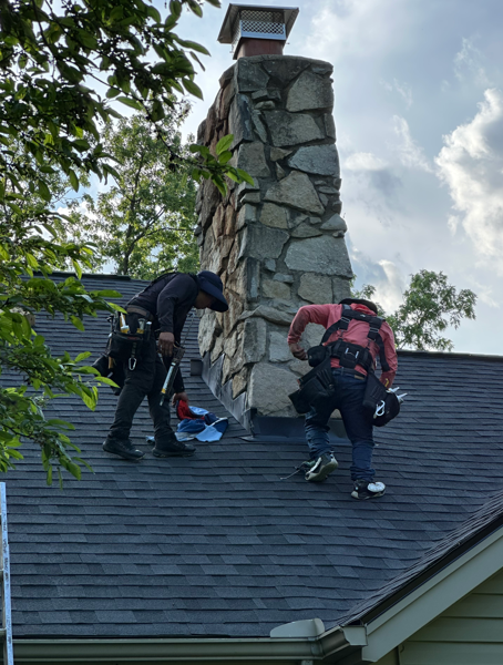 Two roofers repair a shingled roof beside a stone chimney under a partly cloudy sky.
