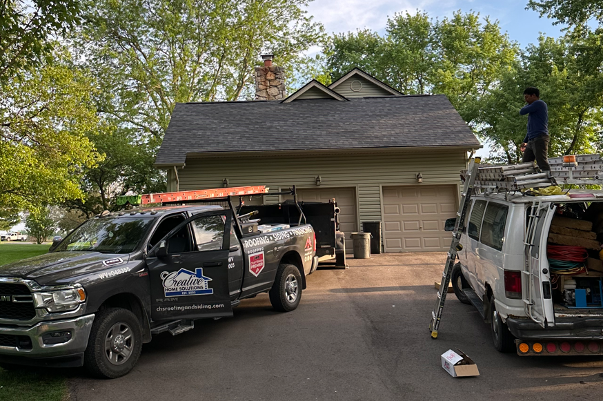 Two utility trucks parked in a driveway beside a house, one worker on the roof with tools.