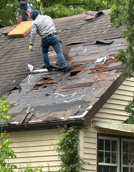Worker repairing a damaged shingled roof on a house, with exposed underlayment and debris
