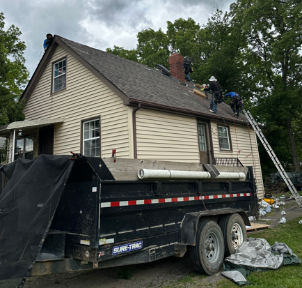 House roof being repaired by workers beside a black dump trailer on a residential street