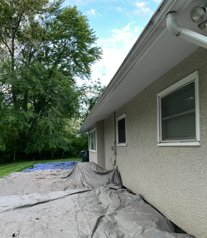 Gray house exterior with roof gutters and windows, covered by tarps along a gravel and tree-lined side yard