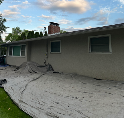 House exterior with gray stucco walls, windows, a brick chimney, and tarps covering the ground during yard work