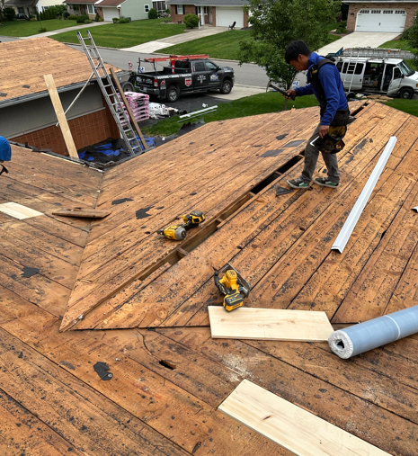 Worker roofing a house, standing on a sloped wooden roof with tools and lumber nearby.