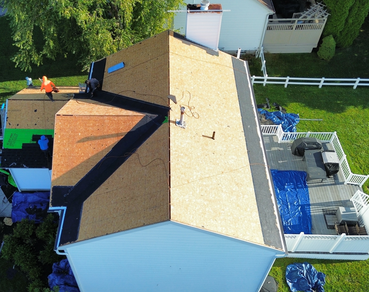 Aerial view of a house roof with workers, tarps, and fencing in a backyard.