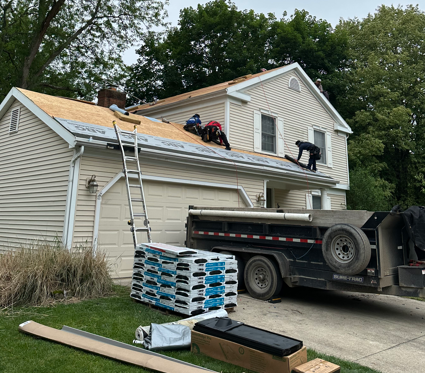 Roofers installing plywood on a house roof beside a ladder and dump truck with shingles.