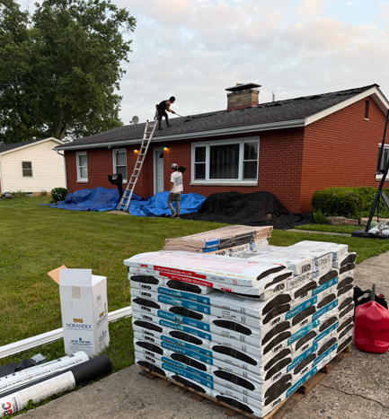 Workers repairing a red brick house roof, with shingles and supplies stacked in the driveway.