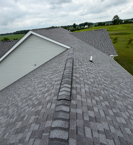 Gray shingled roof with ridge vent on a house, overlooking green fields under cloudy sky