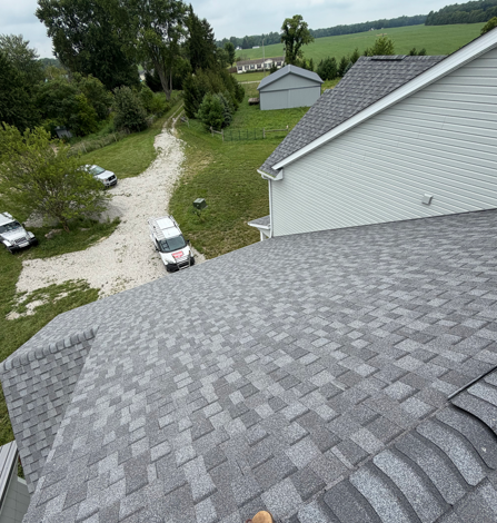 Roof view over a gravel driveway with parked vehicles, trees, and a small outbuilding in the distance