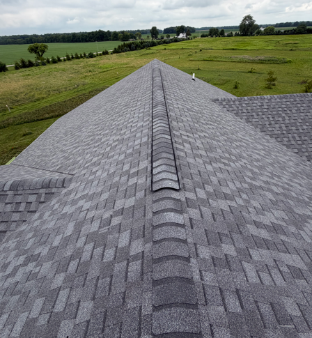 Gray shingled roof ridge overlooking green farmland under a cloudy sky
