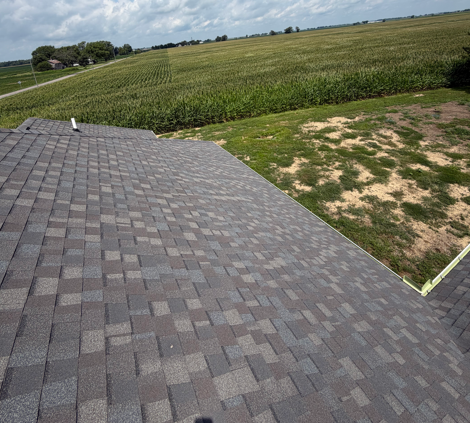 Rooftop with gray shingles overlooking green fields and a small patchy yard under a cloudy sky