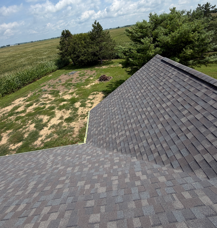 Gray shingled roof overlooking a grassy yard, trees, and open fields under a cloudy sky
