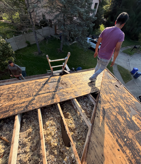 Worker on a steep roof during repairs, with exposed wood and a ladder below.